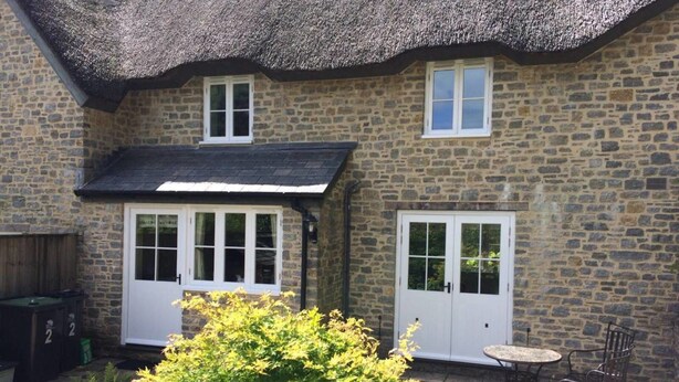A stone house with a thatched roof, two white doors, and a small patio with a table and chairs. - Thatched Cottage, West Dorset (Dorchester)