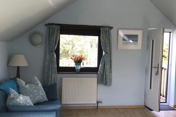 A cozy room with a sloped ceiling, a radiator, a blue sofa with patterned cushions, a window with curtains, a wall clock, and a framed picture.