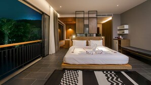 A modern hotel room with a large bed, a balcony with a view of trees, a wooden headboard, and a small desk with a lamp. - The Gallery Hotel Nai Harn (Rawai)
