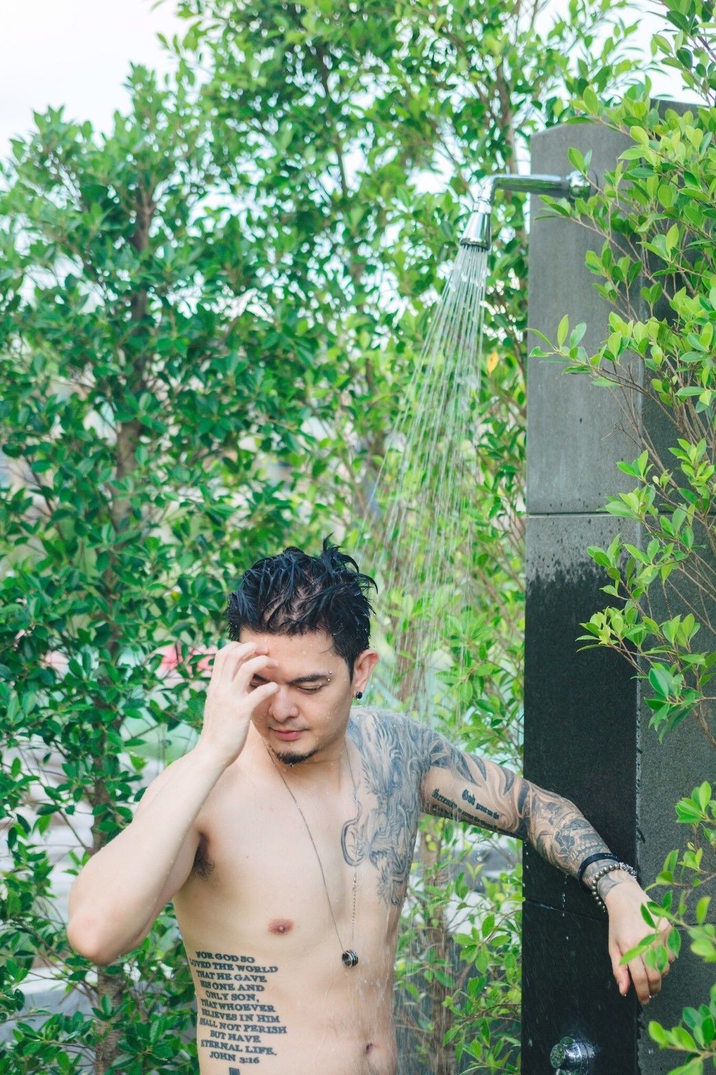A person with tattoos standing under a shower in an outdoor setting with greenery.