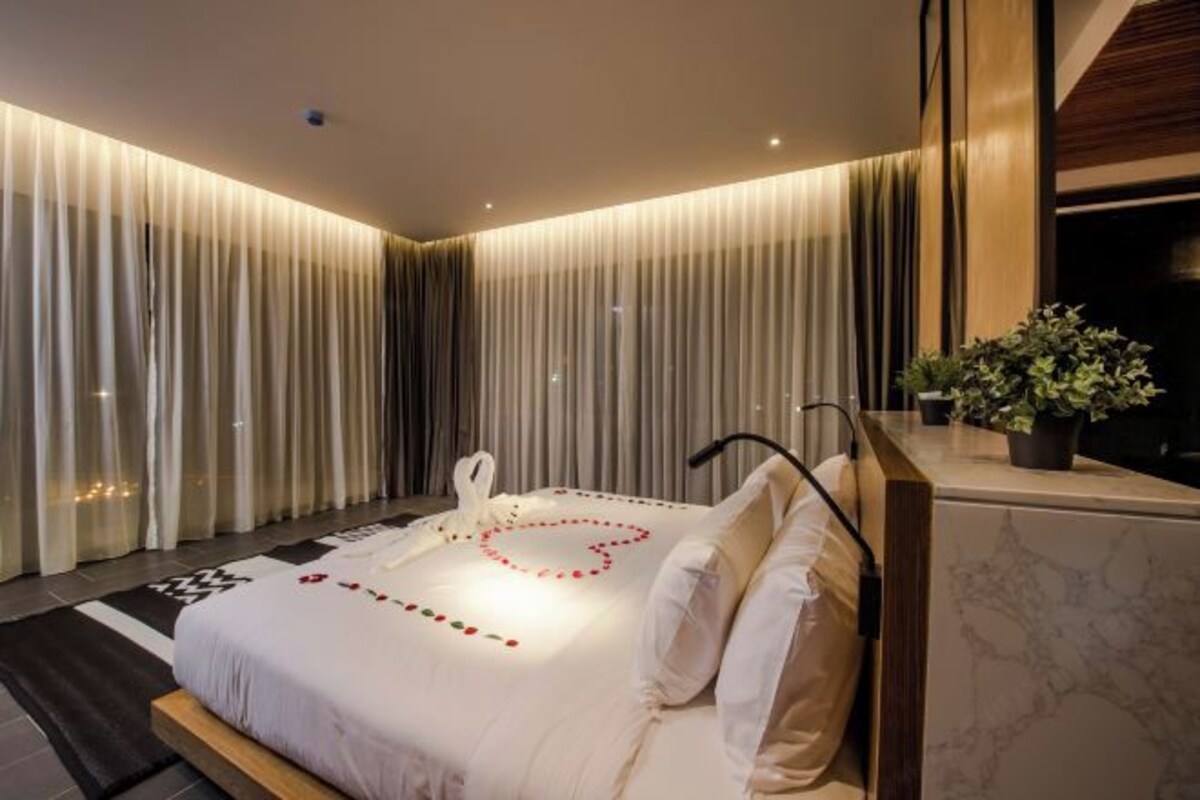 A modern hotel room with a large bed, a heart-shaped decoration on the bed, a small plant on the nightstand, and a view of the city through the window.