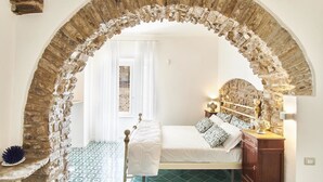 A bedroom with a stone archway, a bed with patterned bedding, a nightstand with a lamp, and a view of the outside. - Palazzo Gentilcore (Castellabate)
