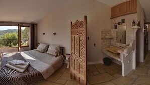 A bedroom with a bed, a TV, a bathroom with a mirror and sink, and a view of the outdoors. - Les Gites du Lac - Chambre d'hotes (Montauroux)