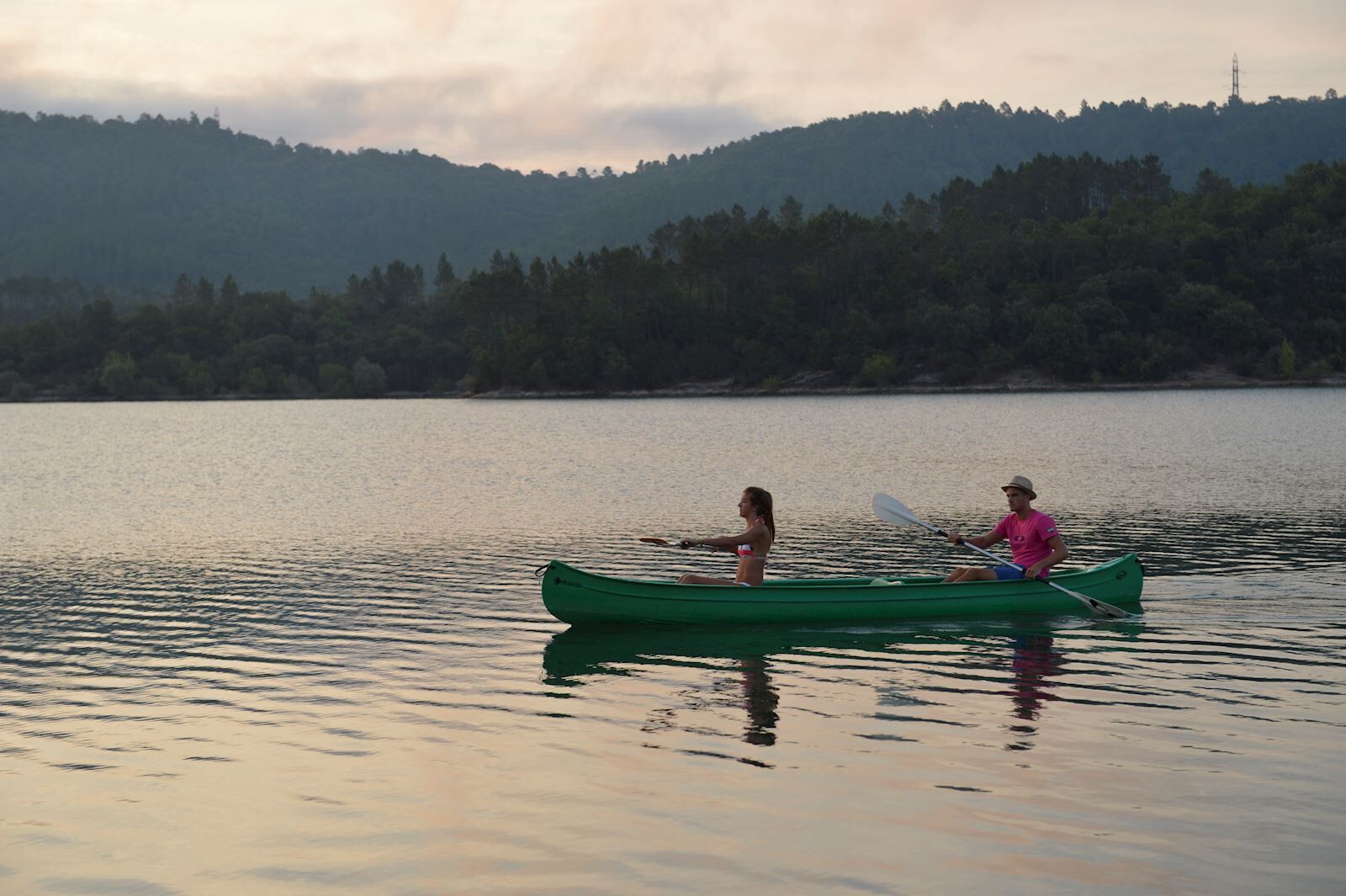 Two people in a green canoe on a calm lake with a forested hill in the background.