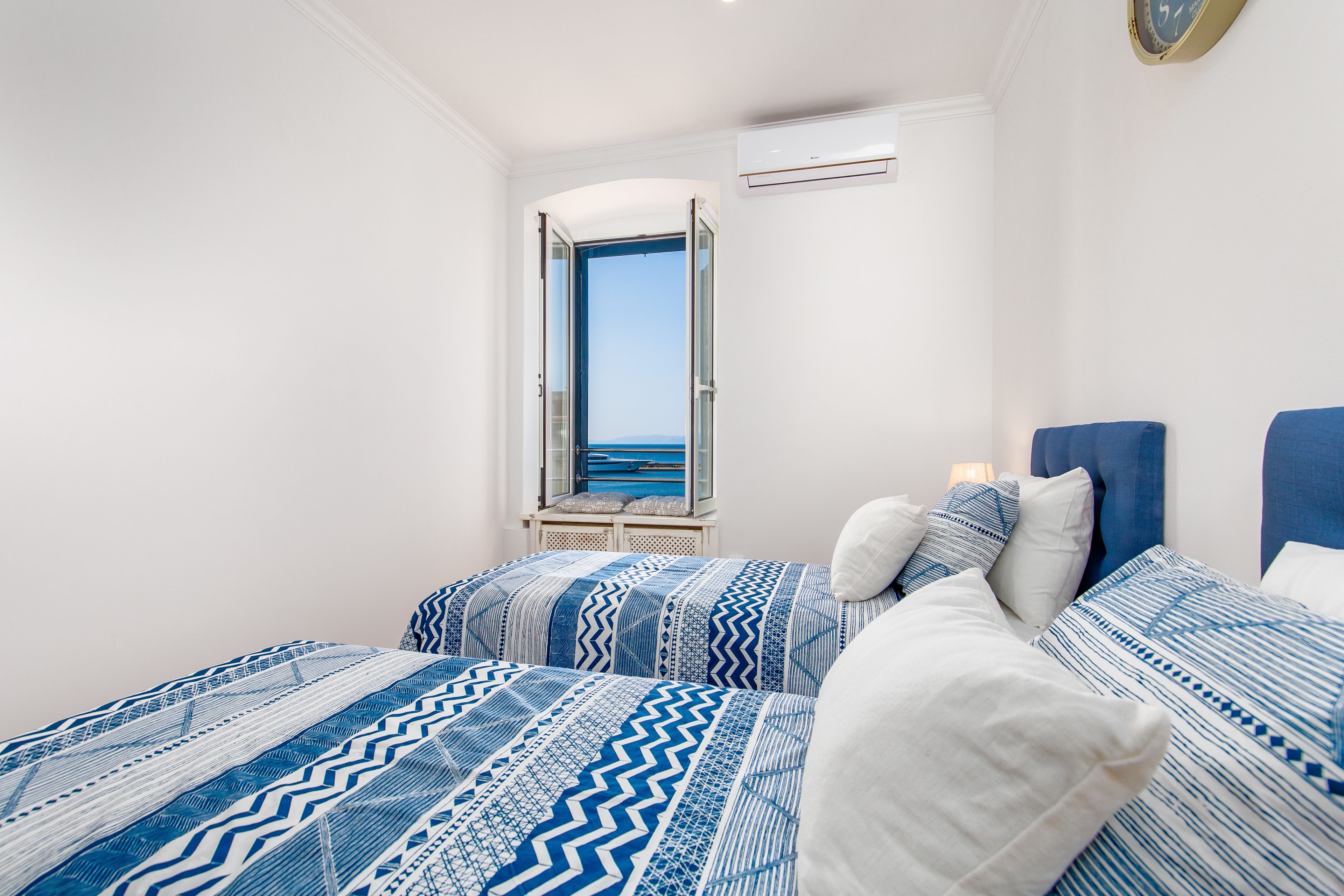 A bedroom with two beds, a window with a view of the sea, and an air conditioning unit on the wall.