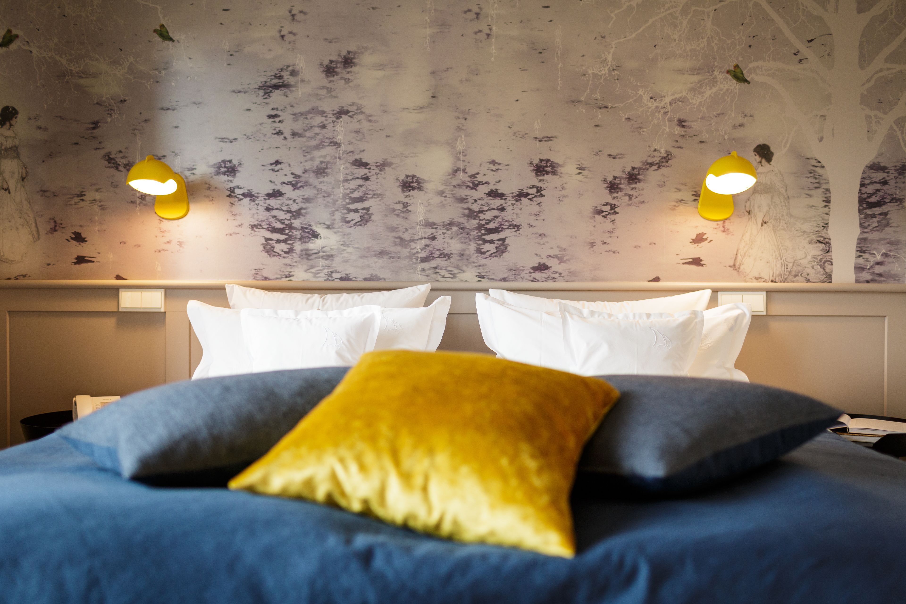 A hotel room with a bed, white pillows, a blue and yellow accent pillow, and a wall mural with tree designs.