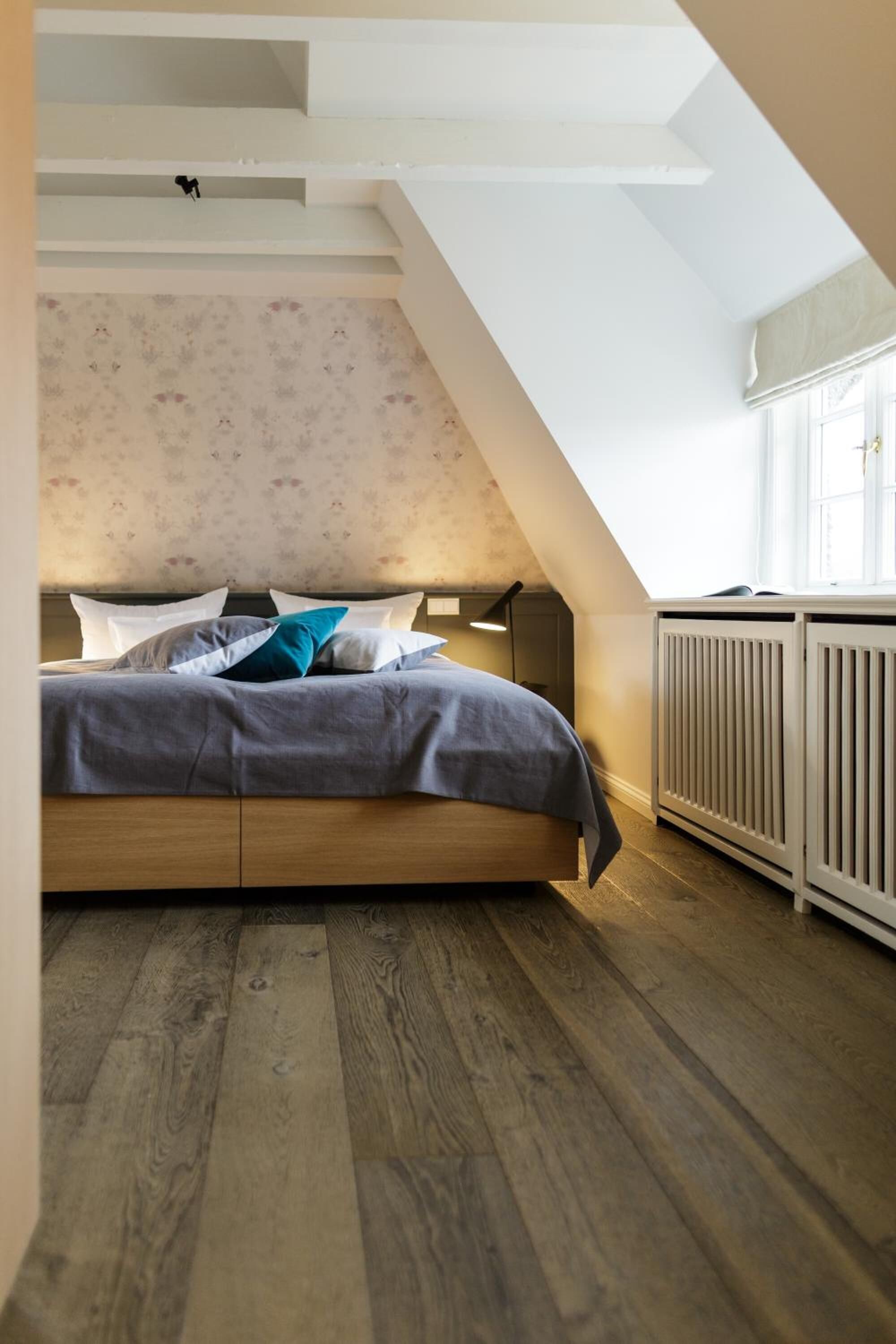 A bedroom with a sloped ceiling, a bed with a wooden headboard, a radiator, and a window with curtains.
