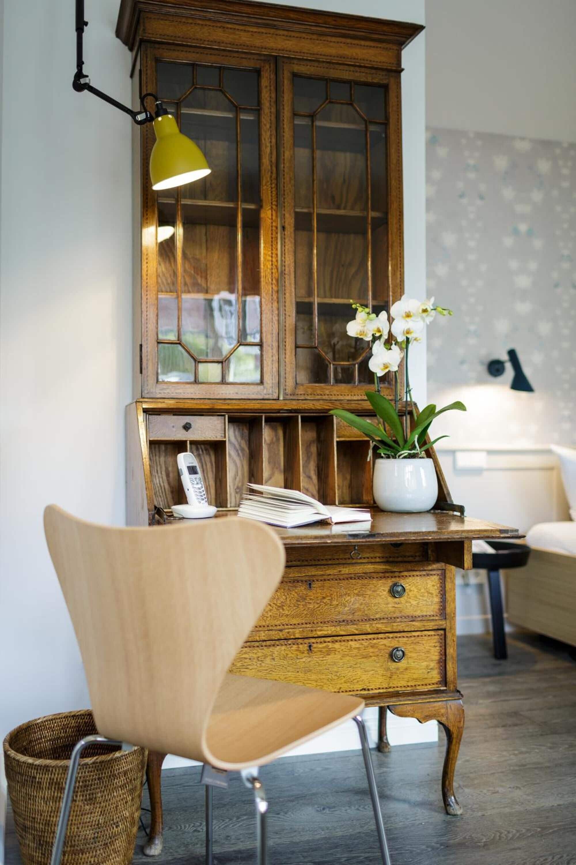 Een houten kast met glazen deuren, een witte telefoon, een orchidee in een pot en een gele bureaulamp.