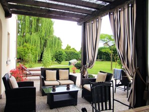 Ein überdachter Patio-Bereich mit Korbmöbeln, darunter eine Couch und Stühle, sowie ein Glastisch.