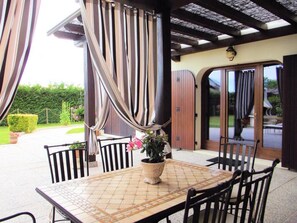 A covered patio with a tiled table and chairs, a potted plant, and a wooden door.