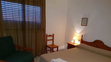Une chambre d’hôtel avec un lit, une chaise, une table de chevet avec une lampe et une fenêtre avec des stores.