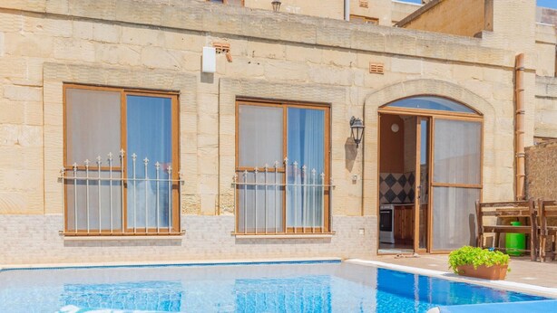 A pool area with a lounge chair and a decorative pillow, a stone building with large windows and a door, and a small table with a plant. - Ta Danjela Villa Sleeps 8 With Pool (Xaghra)
