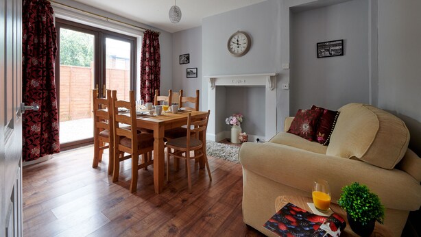 Un salon avec un canapé beige, une table à manger avec des chaises, une cheminée et une horloge au mur.