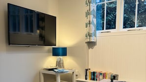Ein Zimmer mit an der Wand befestigtem Fernseher, einem kleinen Tisch mit Lampe, einem Bücherregal und einem Fenster mit Vorhängen.