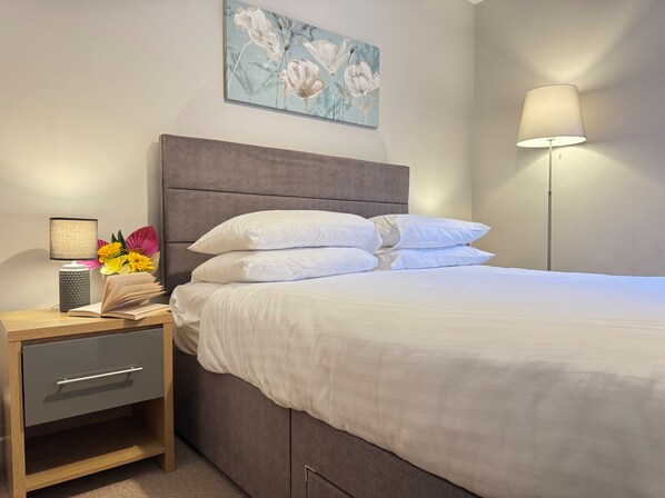 Un letto rifatto con lenzuola bianche, un comodino con una lampada e una piccola pianta decorativa.
