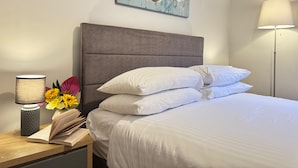 Ein ordentlich bezogenes Bett mit weißen Leinentüchern, einem grauen gepolsterten Kopfteil und einem Nachttisch mit Lampe und Buch.