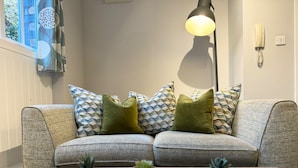 Ein Wohnzimmer mit grauem Sofa, gemusterten Kissen, einer Stehlampe, einem an der Wand montierten Telefon und drei kleinen Topfpflanzen auf einem Holztisch.