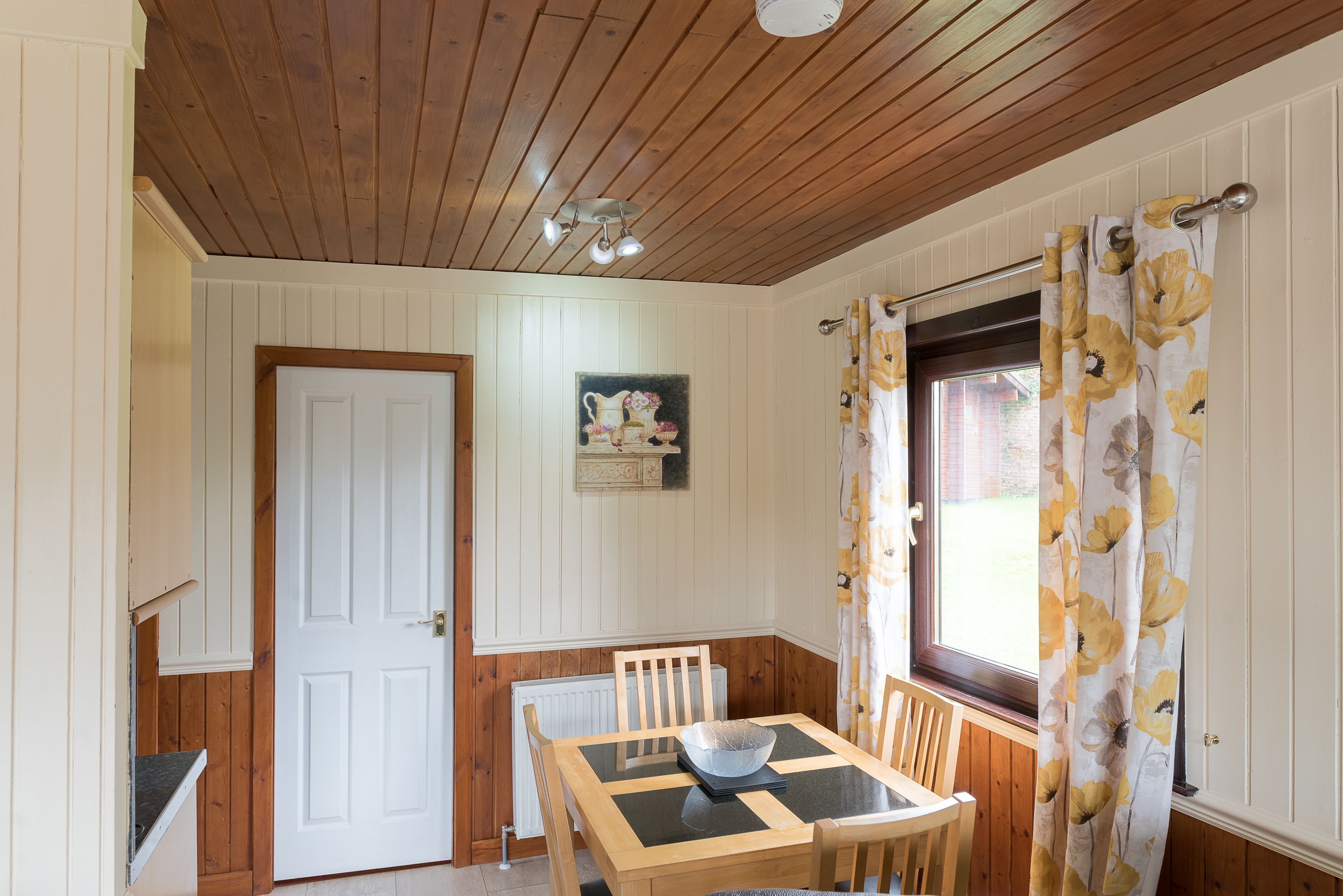 A dining area with a wooden table, chairs, a white door, and a window with floral curtains.