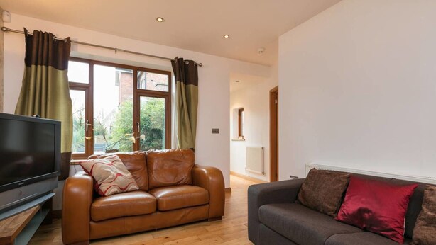 A living room with a brown leather sofa, a grey sofa with red cushions, a flat-screen TV, wooden flooring, and a window with curtains. - Luxury 1bedroom Lodge in Prestwich (Manchester)