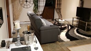 Un salon avec un canapé gris, un téléviseur, une petite table sur laquelle se trouve une bouteille de champagne et une horloge décorative accrochée au mur.