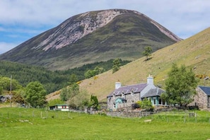 A stone house with a sloped roof and a chimney, surrounded by a fenced field and a mountain in the background.