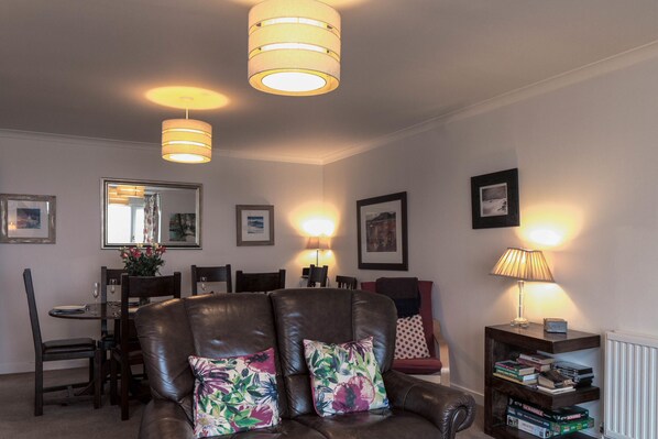 Un salon avec un canapé en cuir marron, deux coussins à motifs floraux, une table à manger avec des chaises, un miroir et des lampes fixées au mur.