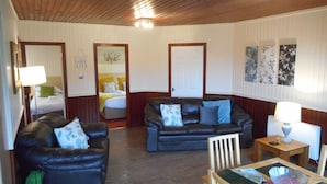 Una habitación con dos sofás de cuero, una mesa de comedor de madera y una silla. Se ve una cama con cabecera blanca y una puerta con marco blanco.