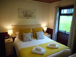A neatly made bed with a yellow bedspread, two white towels, and patterned pillows. There are two bedside lamps, a nightstand with a phone, and a window with curtains.