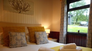 Una cama bien hecha con colcha amarilla y toallas blancas, una mesita de noche con lámpara, y una ventana que da a una casa y un coche.