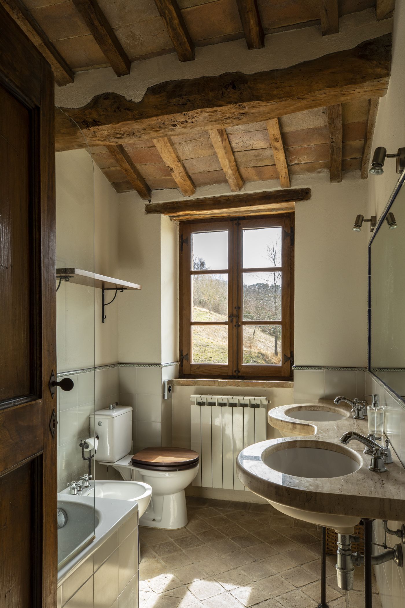 A rustic bathroom with wooden beams, a window, a toilet, and two sinks.