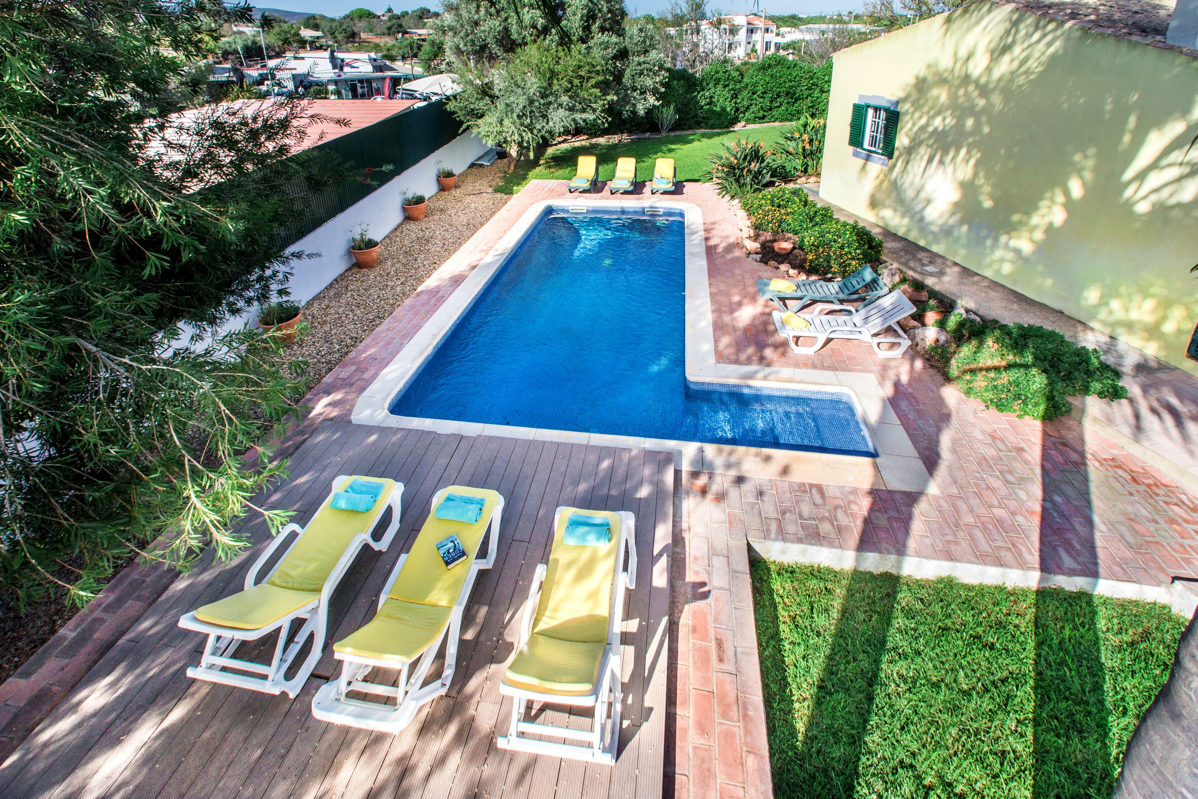 A rectangular swimming pool with a tiled deck, surrounded by potted plants and a grassy area.