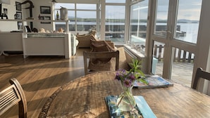 Amplia sala de estar con un gran ventanal con vista al agua, una mesa de comedor con un florero y una zona de estar acogedora con muebles de mimbre.