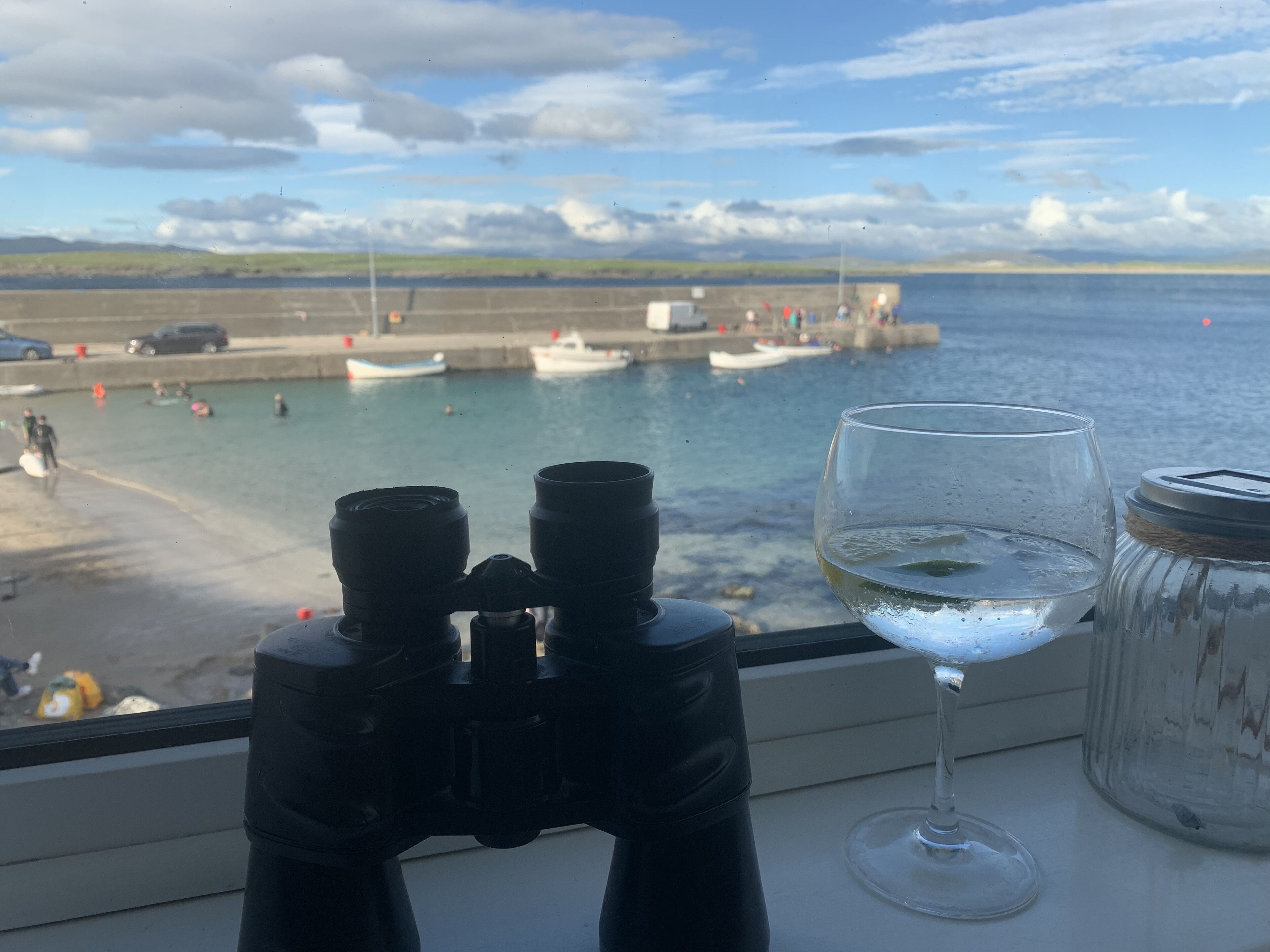 Binoculars and a glass of water on a windowsill with a view of a harbor and boats.