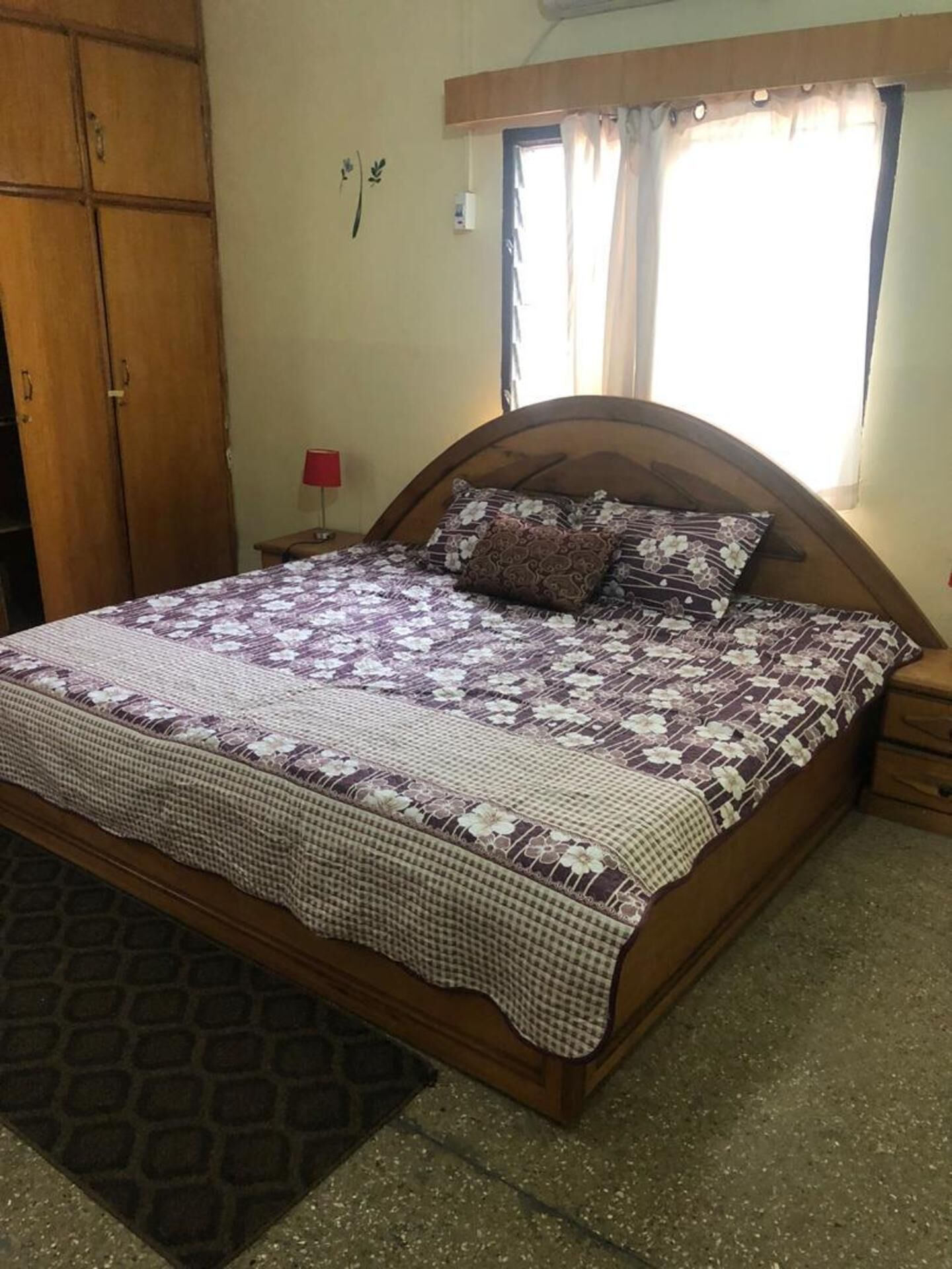 A bedroom with a wooden bed, a checkered bedspread, a nightstand with a lamp, and a window with curtains.