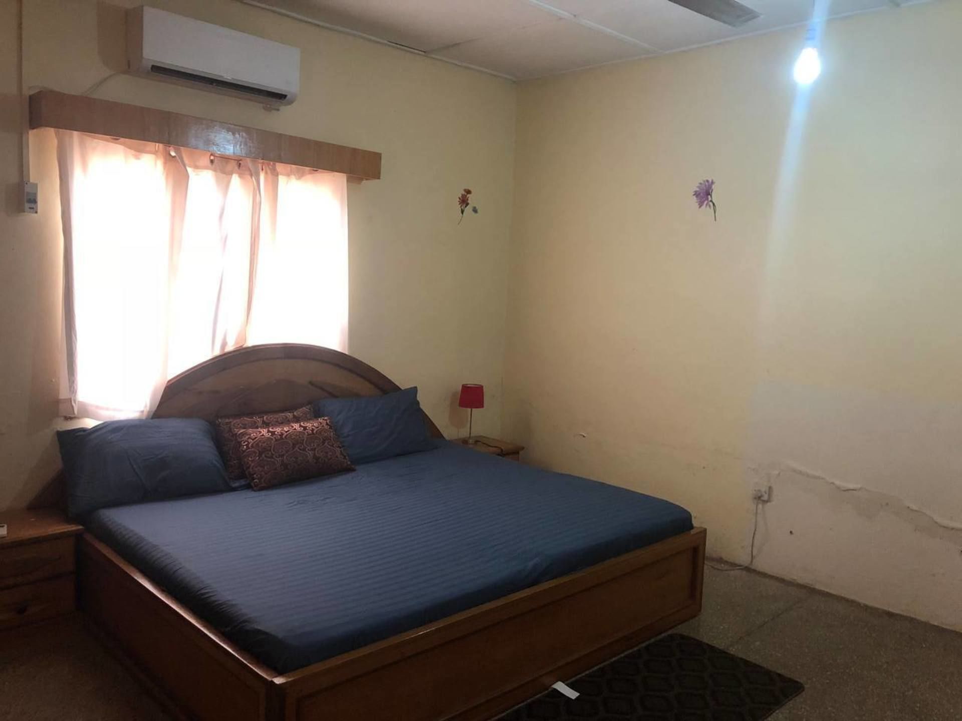 A single-bed room with a wooden headboard, a bedside table, a red lamp, and a wall-mounted air conditioner.