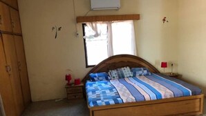 A bedroom with a wooden bed, a nightstand, a red lamp, a window with curtains, and an air conditioning unit. - 3 Bed House Sleeps 6 - Parking, Wifi, Generator (Kumasi)
