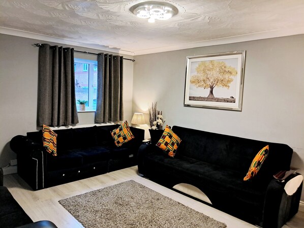 A living room with a black sofa set, a grey rug, a framed tree painting, and a window with curtains. - London Excel 3bed House Sleeps 6 - Car Park for 2 (London)