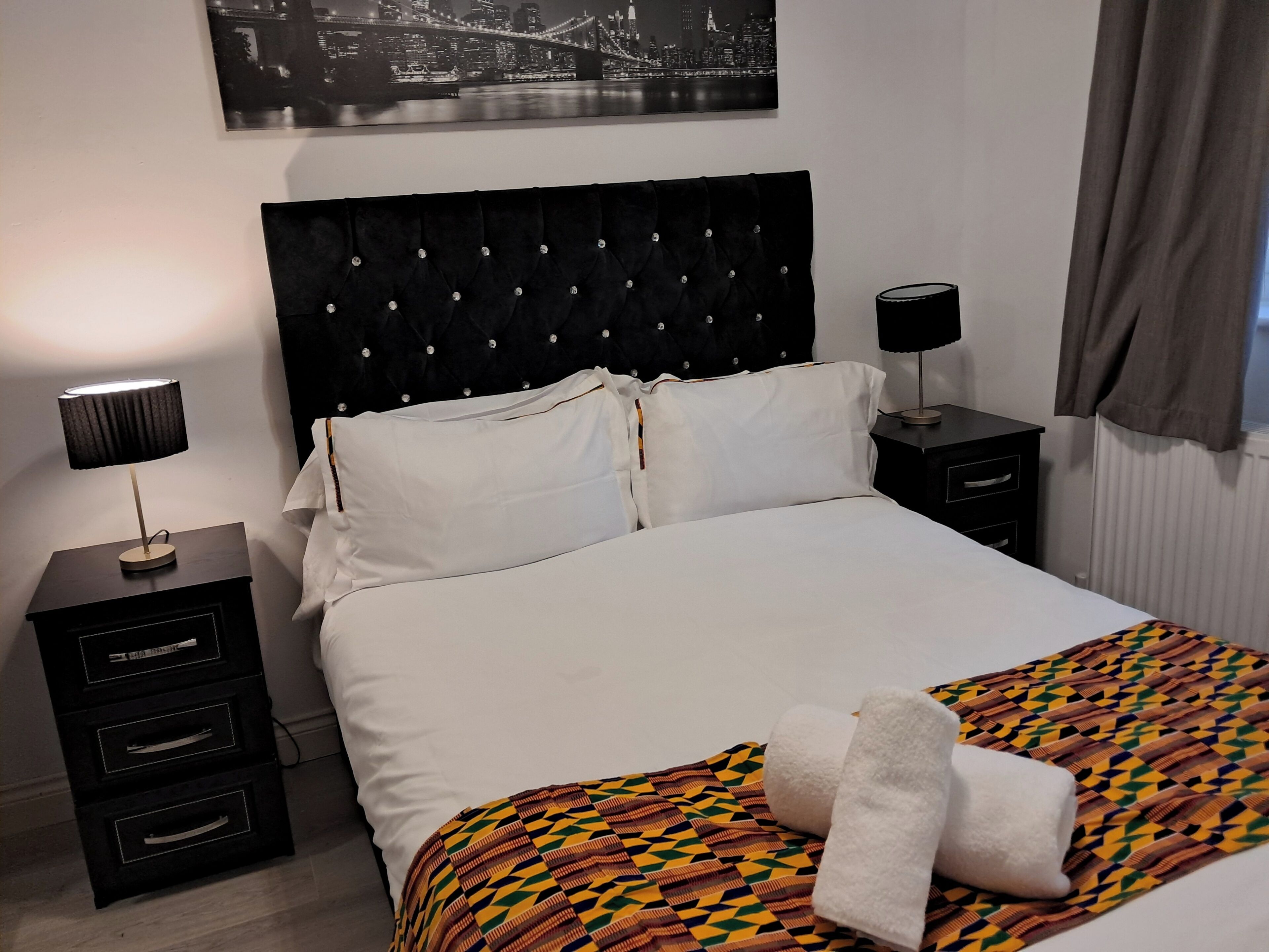 Een net opgemaakt bed met witte lakens, een zwart hoofdbord en een kleurrijke geborduurde deken. Er staan twee nachtkastjes met lampen en een kleine ladekast bij.