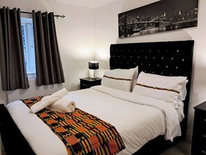 A neatly made bed with white linens and a colorful blanket, a black headboard, a nightstand with a lamp, and a cityscape painting on the wall. - London Excel 3bed House Sleeps 6 - Car Park for 2 (London)