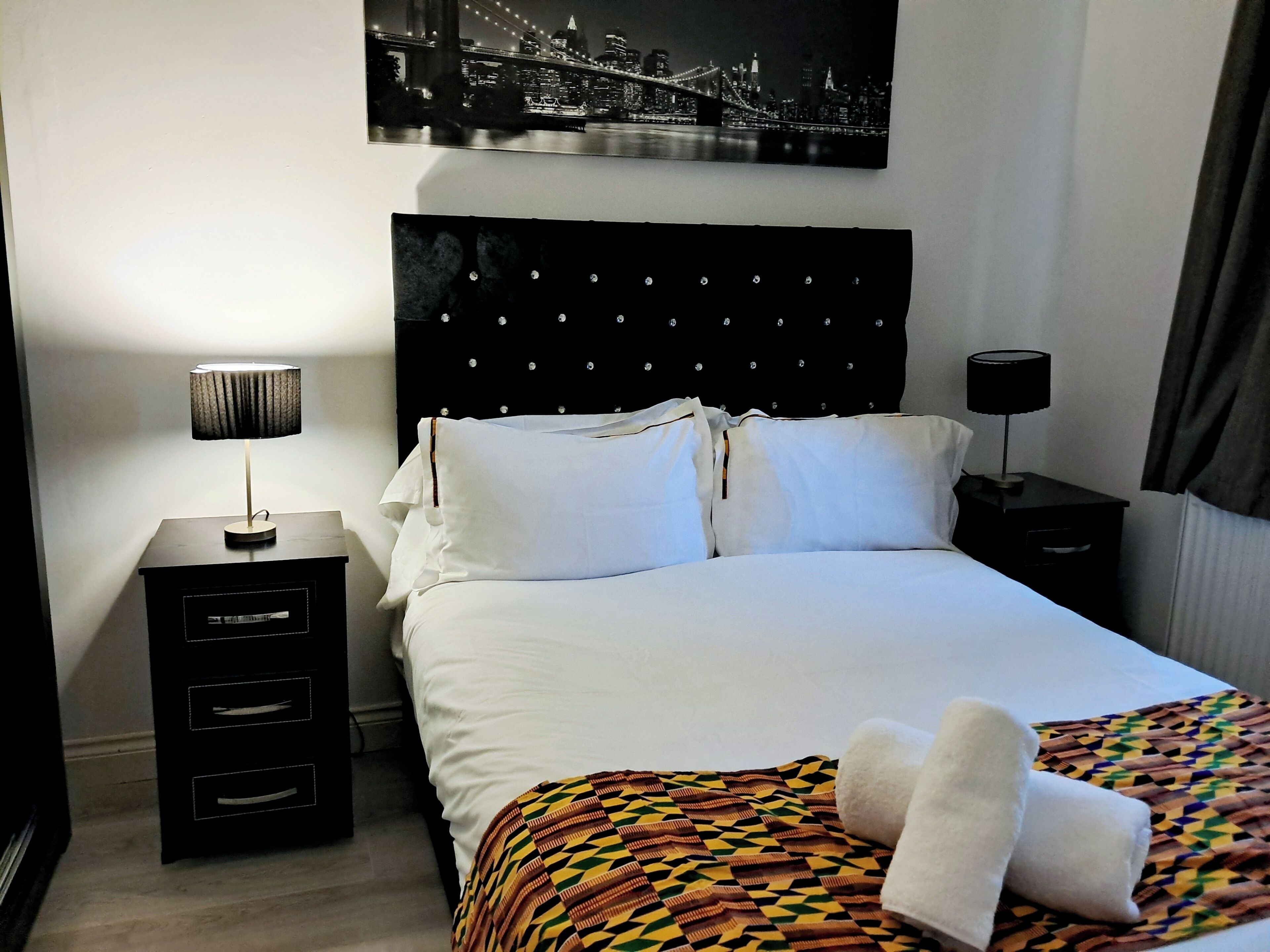 A hotel room with a neatly made bed, a black headboard with a cityscape photo, a side table with a lamp, and a patterned throw blanket.