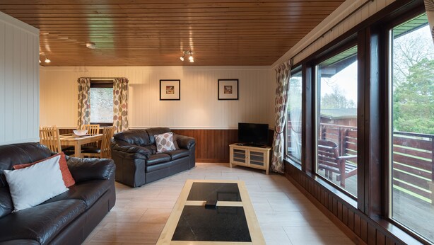 Una sala de estar acogedora con un sofá de cuero, una mesa de centro y un mueble para televisión. Amplias ventanas ofrecen vistas al exterior.