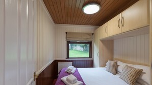 Un dormitorio con techo de madera, una ventana con vistas a zonas verdes, una cama con sábanas blancas y ropa de cama morada, y un radiador debajo de la ventana.