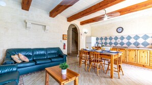 A spacious living area with a dark green sofa, wooden dining table, and chairs. A small wooden table with a plant is in the foreground. The room features exposed wooden beams, a tiled backsplash, and a kitchen area with wooden cabinets and a clock on the wall. - Villa Ta Debora Sleeps 7 With Pool (Xaghra)