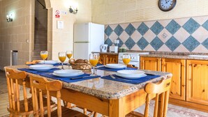 A kitchen with a granite countertop, wooden chairs, a dining table set with plates and glasses, a refrigerator, a stove, and a wall clock. - Villa Ta Debora Sleeps 7 With Pool (Xaghra)