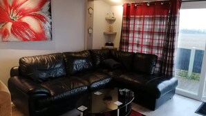 A living room with a black leather sofa, a glass coffee table, a red and black rug, a large abstract painting, a red plaid curtain, and a small shelf with decorative items. - Sea View Retreat - Free Wifi (Bridlington)