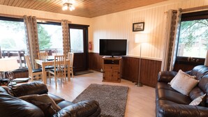 A living room with a leather sofa, a TV on a wooden stand, a dining table with chairs, and large windows with curtains. - Birch Lodge 23 With Hot Tub, Newton Stewart (Newton Stewart)