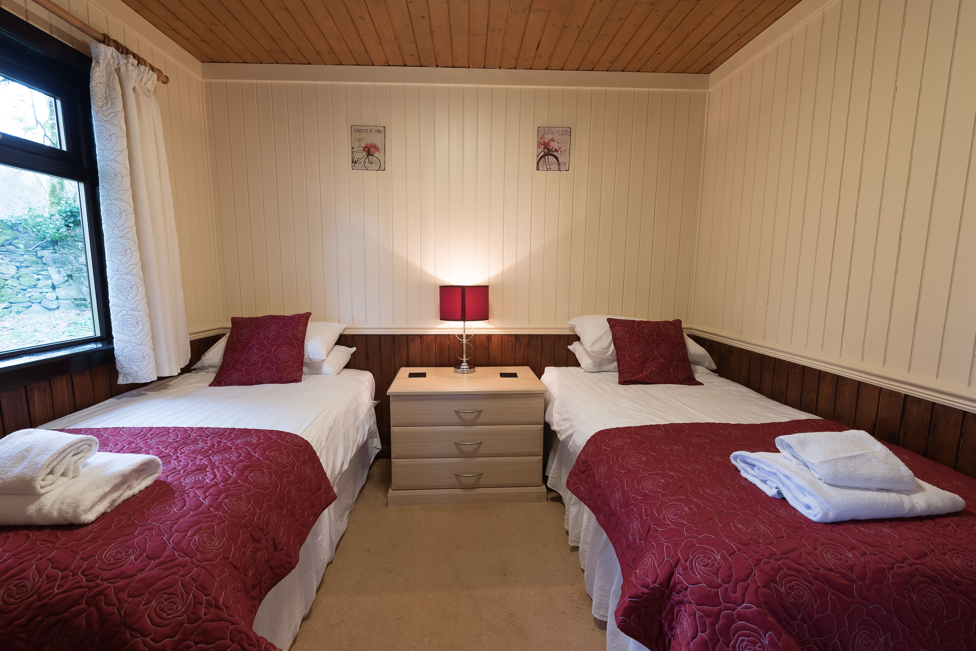 Habitación doble con dos camas, cada una con una colcha roja, una mesita de noche de madera, una lámpara y una ventana con cortinas.