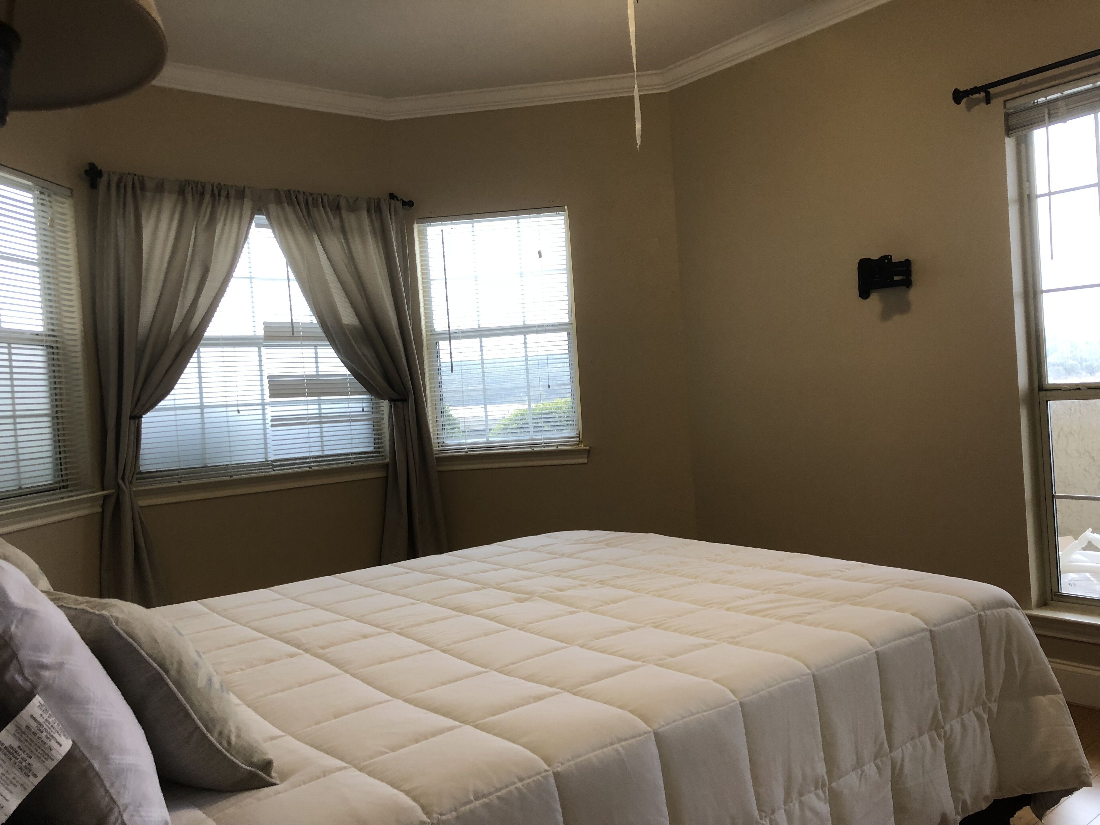 A bedroom with a bed, two windows with curtains, a ceiling fan, and a thermostat on the wall.
