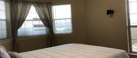 A bedroom with a bed, two windows with curtains, a ceiling fan, and a thermostat on the wall.