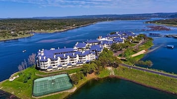 Une vue aérienne d’un grand hôtel à plusieurs étages, doté d’un court de tennis et situé près d’une étendue d’eau.
